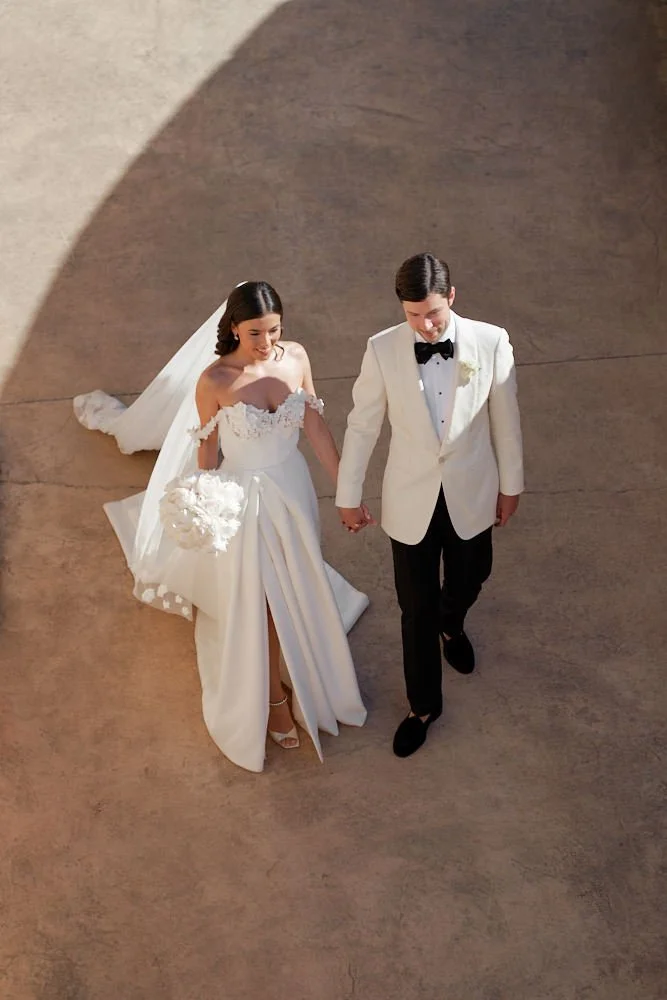 A bride and groom holding hands, walking on a concrete surface, with the bride in a white off-shoulder wedding gown holding a bouquet and the groom in a white tuxedo jacket, black pants, and a black bow tie.