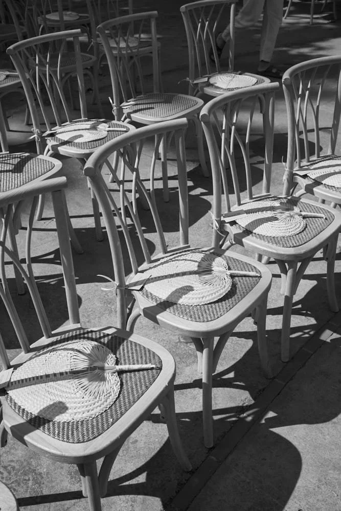 Multiple chairs with woven fans placed on their seats, arranged outdoors on a concrete surface, with shadows cast by sunlight.