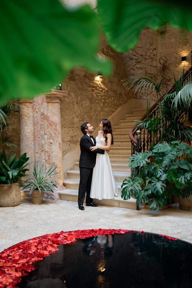 A bride and groom are standing in an indoor garden with lush green plants, stone walls, and a curved staircase, gazing at each other during their wedding or engagement photos.