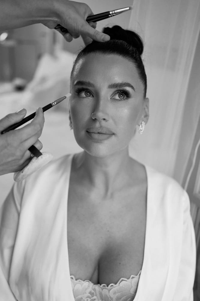 A woman with dark hair styled in a bun is having makeup applied to her face by a makeup artist. The woman is wearing earrings and a white top with a lace detail. The setting appears to be indoors, possibly a salon or a dressing room.