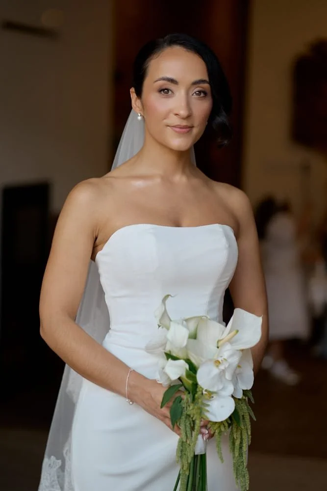 A bride in a strapless white wedding dress holding a bouquet of white calla lilies, with a veil and pearl earrings, standing indoors.