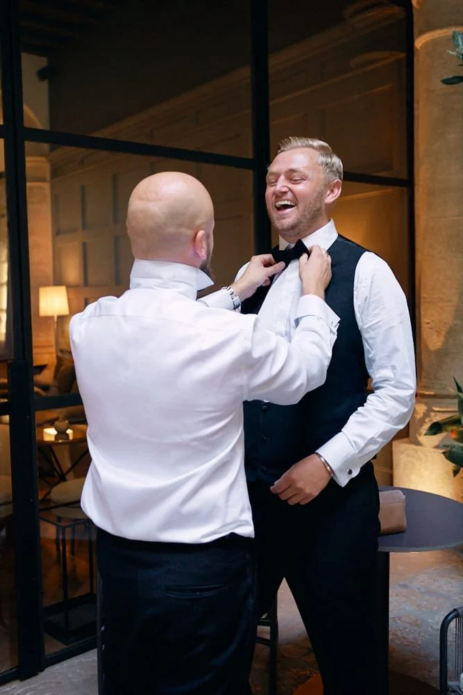 Two men smiling, one helping the other adjust a bow tie at a formal event, with warm lighting and elegant decor in the background.
