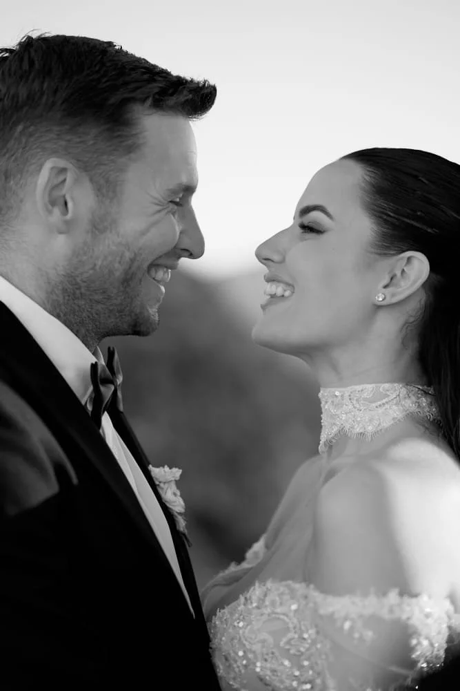 A black and white photo of a smiling couple in wedding attire, facing each other closely.