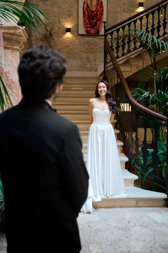 Woman in a white strapless dress smiling on a staircase, man in a black suit in the foreground, inside a building with stone walls and plants.