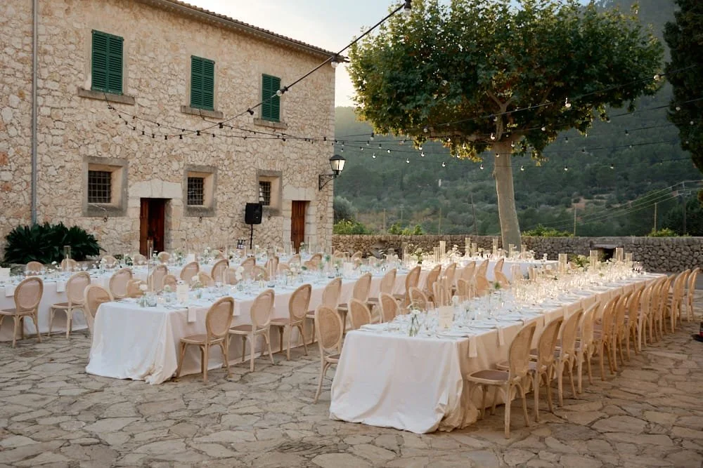 Outdoor dining setup with long tables covered in white tablecloths, set with glasses, plates, and floral centerpieces, under string lights and near a stone wall building and large tree in a scenic outdoor setting.