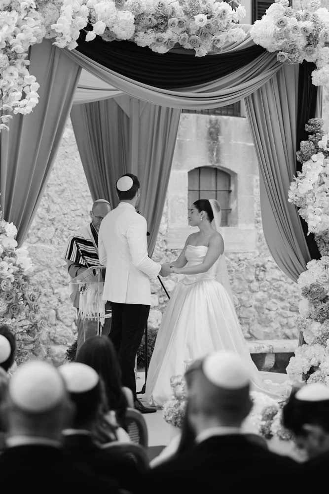 A couple getting married at an outdoor ceremony, standing under a decorated arch with flowers, with guests seated in front and a religious officiant conducting the vows.