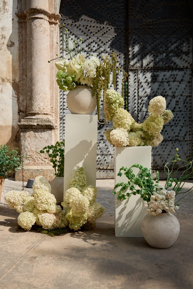 Decorative floral arrangement with white hydrangeas and green foliage in vases on white pedestals outside near an old stone wall and metal gate.