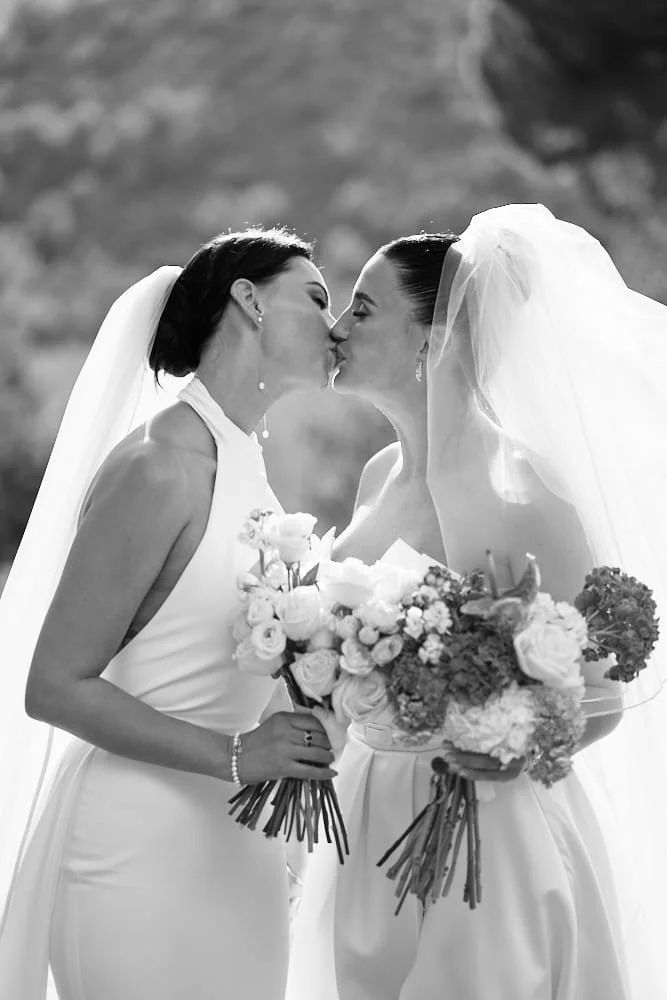 Two women in wedding dresses sharing a kiss, each holding bouquets of flowers, outdoors with a blurred natural background.