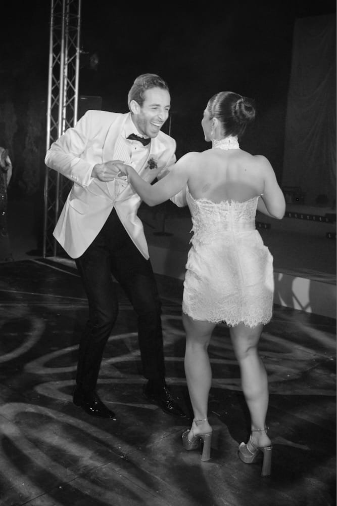 A man and a woman dancing together at an event, with the man wearing a tuxedo and the woman in a short lace dress and high heels.