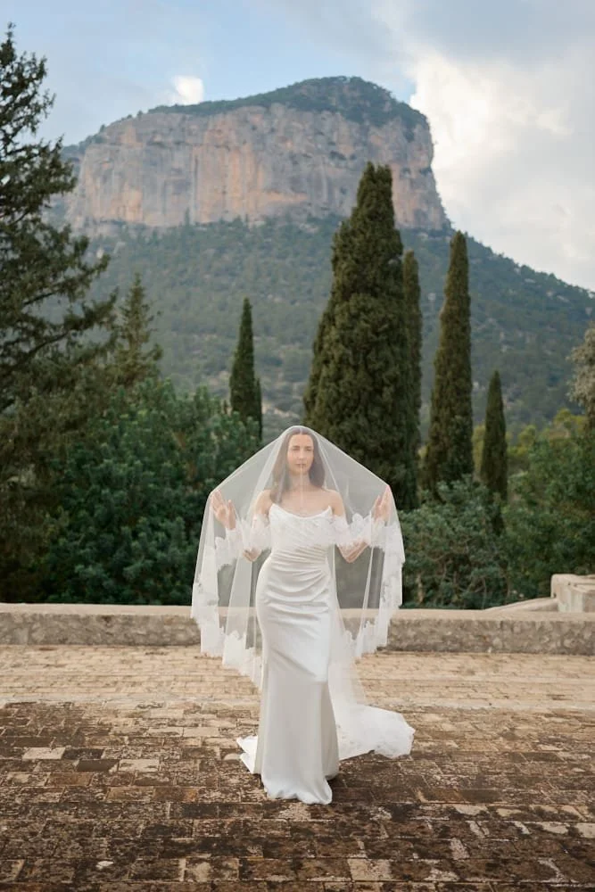 A woman in a white wedding dress standing on a brick terrace surrounded by tall cypress trees with a mountain in the background and a cloudy sky.