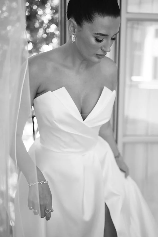A woman in a strapless white dress, likely a wedding gown, looking down with a contemplative expression in a well-lit room.