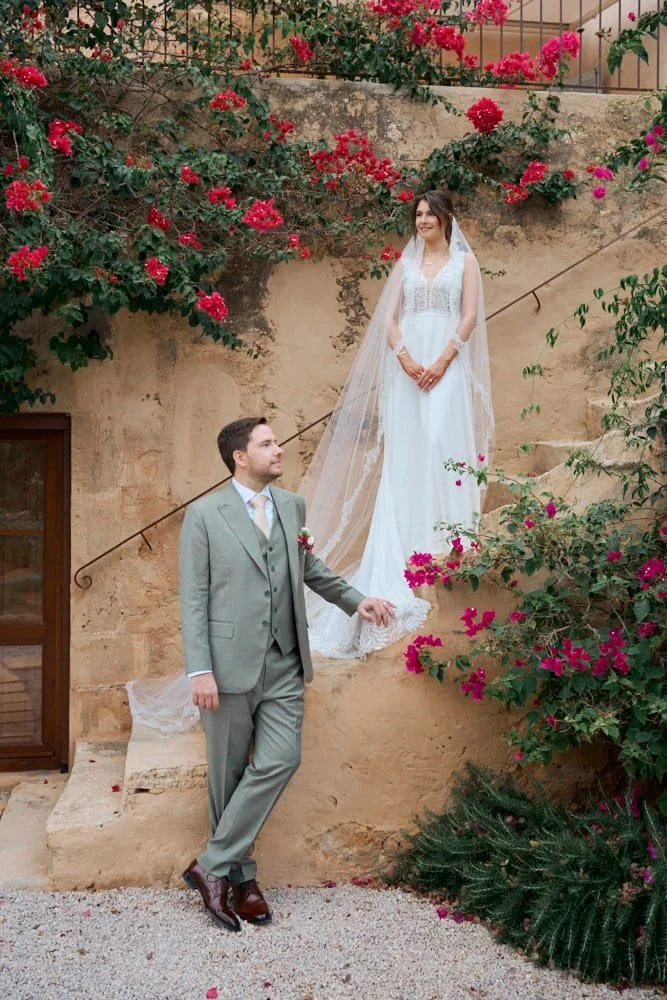 A bride in a white wedding dress with a veil, standing on stairs against a stone wall adorned with pink and red flowers, smiling. A groom in a light gray suit with a boutonnière is standing on the ground, looking up at her.