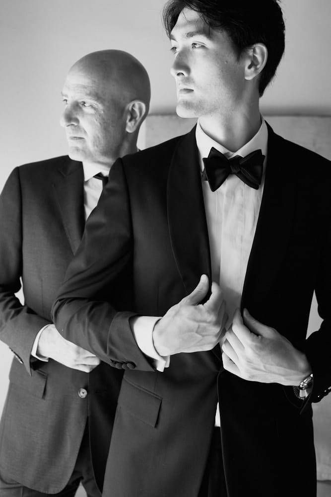 Two men dressed in tuxedos, one bald and the other with dark hair, standing indoors. The man with dark hair is adjusting his jacket while looking to the side. The other man looks forward with a serious expression.