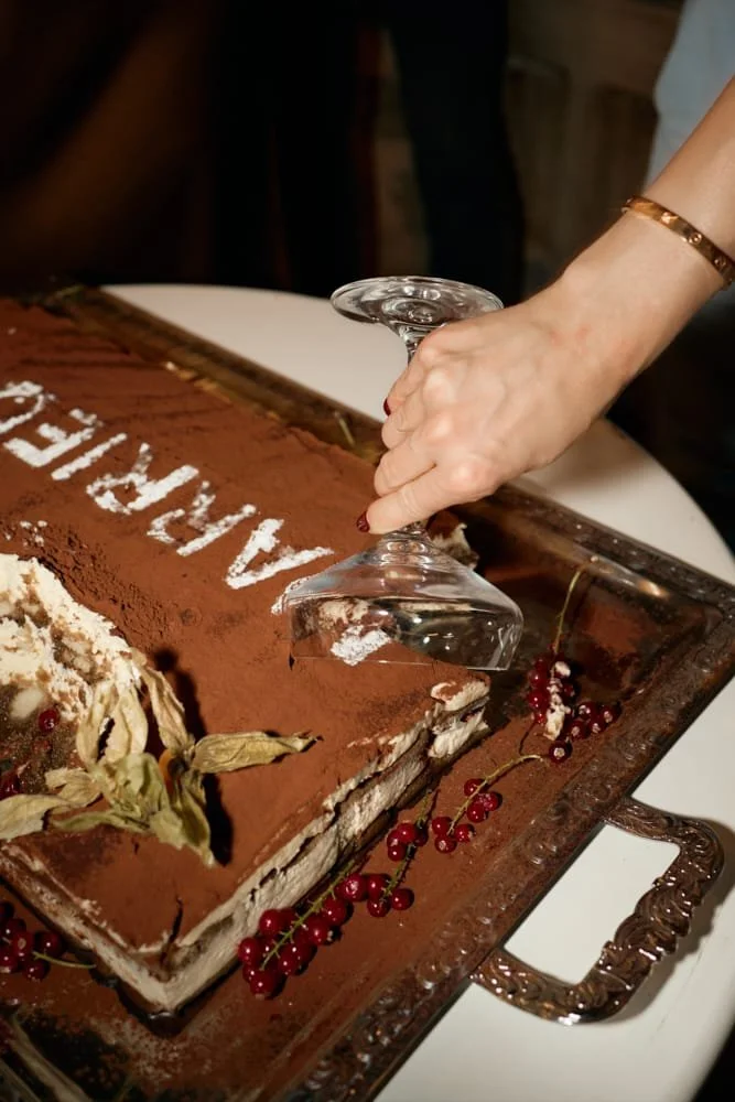 Person placing a wine glass on a rectangular tiramisu cake with cocoa powder and red currants on a decorative tray.