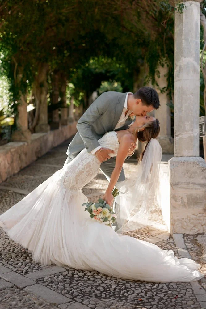 A bride and groom sharing a kiss outdoors; the bride is in a white wedding gown holding a bouquet; the groom is in a gray suit.