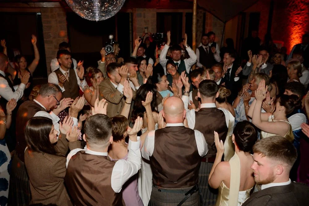 People dancing and celebrating at a wedding reception with a disco ball overhead in a warmly lit venue.