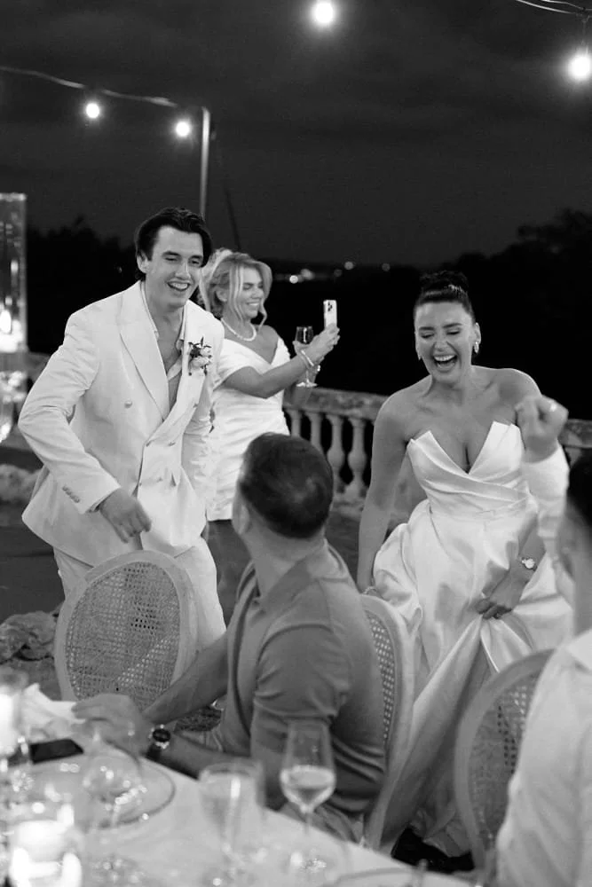 Black and white photo of a wedding reception outdoors at night with string lights overhead. The groom and bride are dancing and smiling, surrounded by seated guests. The bride is in a strapless wedding dress, and the groom is in a light-colored suit.