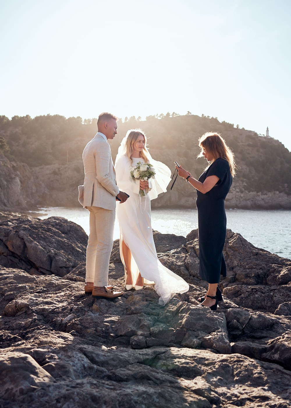 Outdoor wedding ceremony on rocky coastline with officiant, bride, and groom.