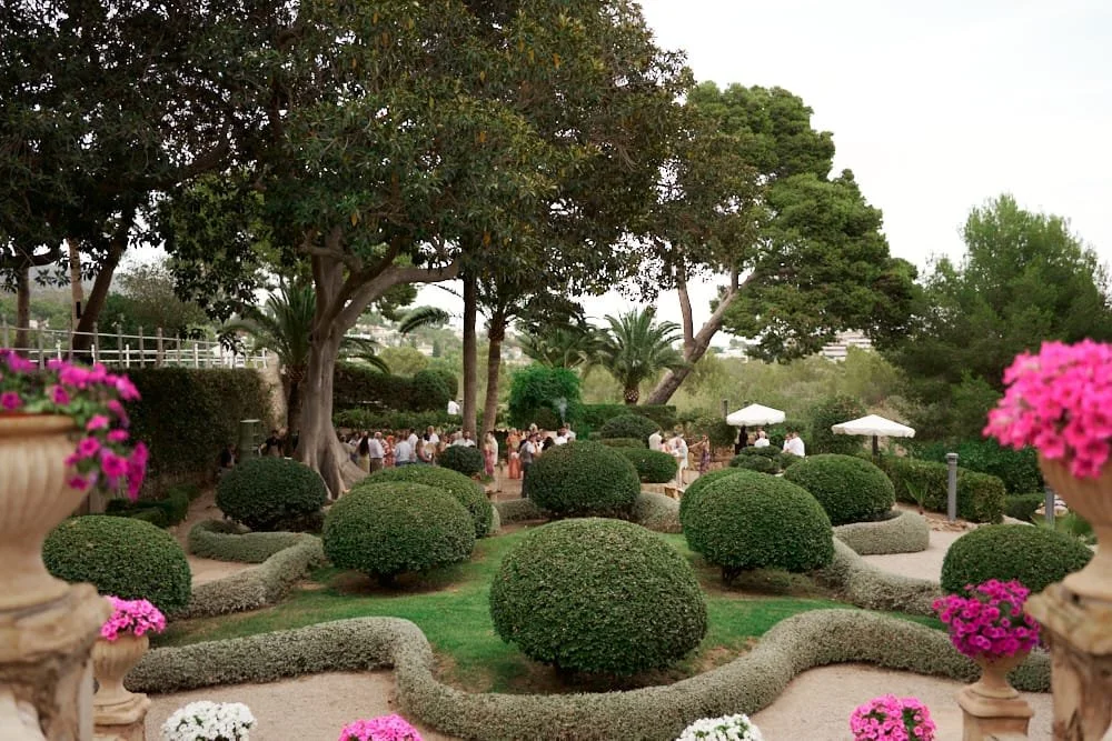 A landscaped garden with large green trees, well-maintained bushes, and pink flowers in decorative pots, with people gathering in the background.