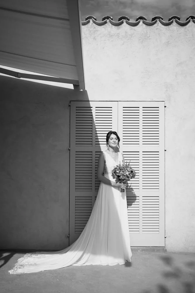 A woman in a wedding dress holding a bouquet standing against a textured wall and decorative shuttered door, with shadow cast on the wall.
