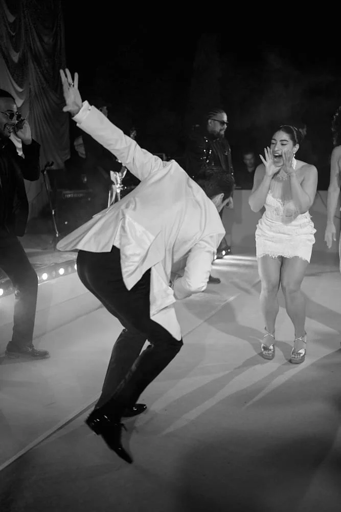 People dancing and celebrating at an event, with a man in a suit jumping and a woman in a white dress smiling.