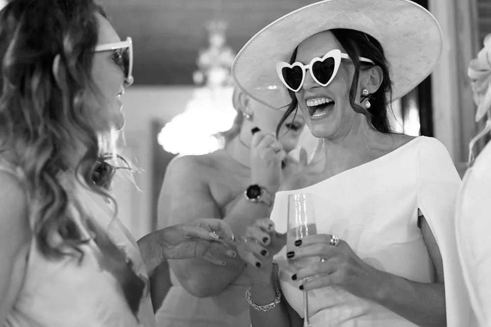 Women at a party wearing stylish outfits and sunglasses, smiling and talking, with one holding a glass of champagne.