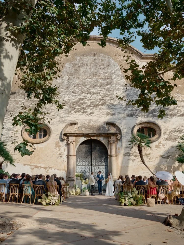 A wedding ceremony taking place outdoors in front of an old, textured stone wall with large wooden doors. The bride and groom are standing at the altar, with guests seated in wooden chairs on either side. Decorative flowers and greenery surround the 