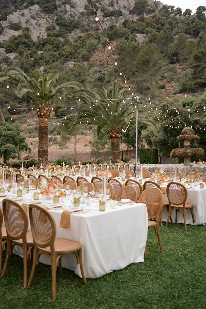 Outdoor event setup with long tables covered in white tablecloths, decorated with floral centerpieces, small vases, and place settings, surrounded by wooden chairs with woven backs, under string lights hanging between palm trees with a mountainous ba