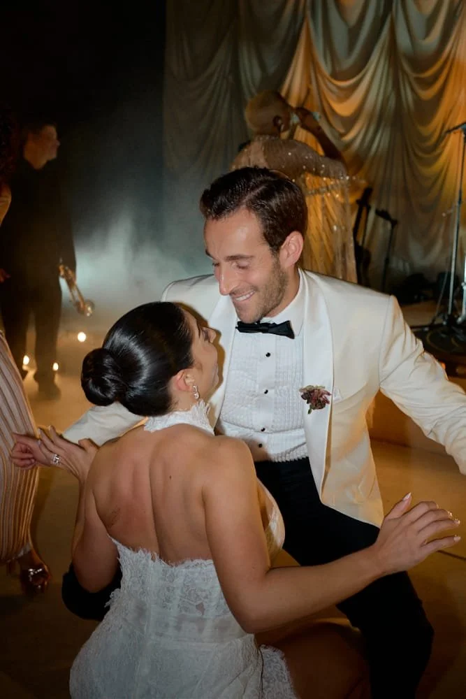 A newlywed couple dancing happily at their wedding reception. The bride is in a white lace wedding dress with her hair in a bun, and the groom wears a white tuxedo jacket with a black bow tie. They are gazing at each other, smiling, with a stage and 