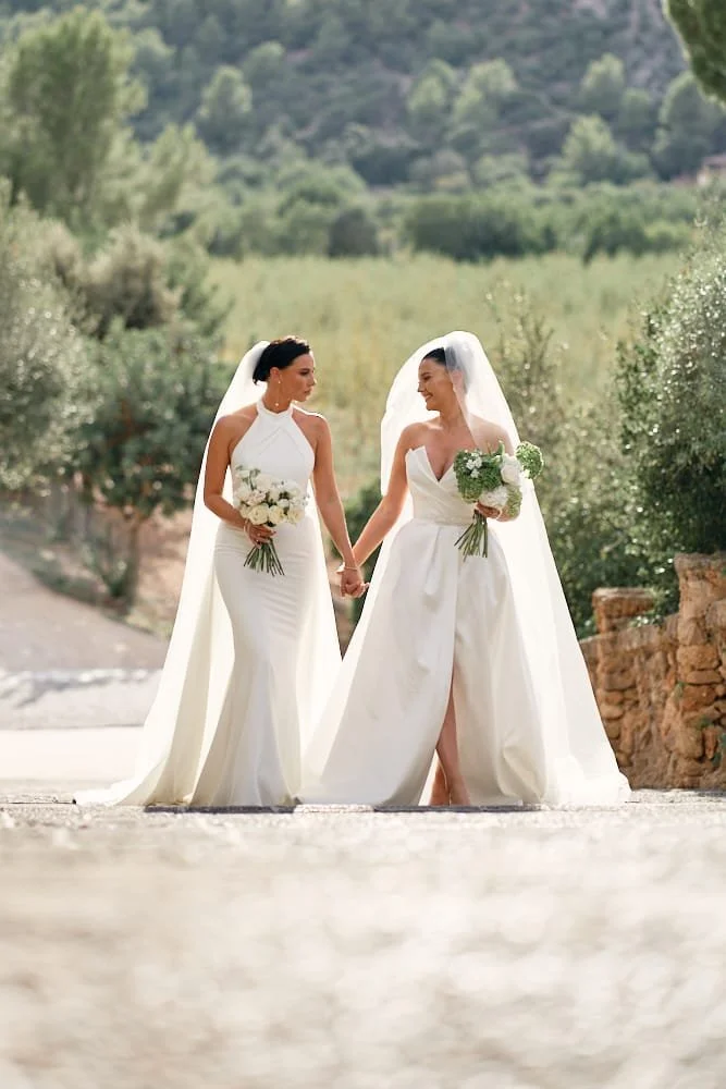 Two women in wedding dresses holding hands and bouquets, walking outdoors with green trees and mountains in the background.