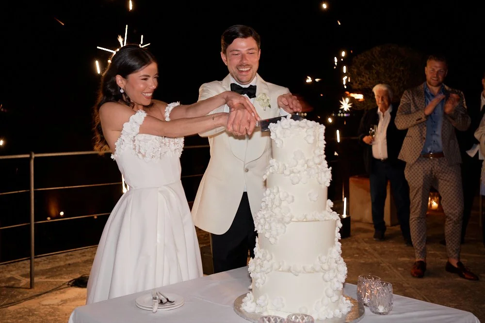 Bride and groom cutting a wedding cake outdoors at night, with guests clapping in the background, fireworks in the sky.