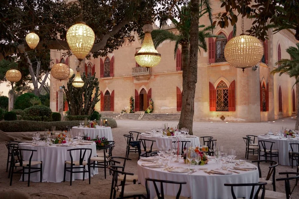 Elegant outdoor dining setup with round tables draped in white tablecloths, decorated with floral centerpieces, under hanging lanterns, with a historic building with arched windows and red shutters in the background.