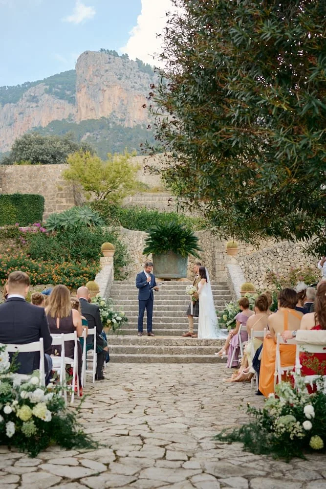 A wedding ceremony outdoors on stone steps with a mountainous landscape in the background, where a groom and bride stand facing each other with an officiant; guests are seated on either side, and the scene is decorated with flowers and greenery.