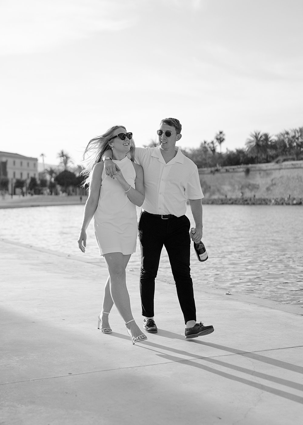Man and woman walking by the water, both wearing sunglasses, man holding a bottle, trees and buildings in background.