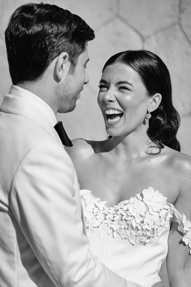A black-and-white photo of a smiling woman wearing a strapless floral wedding dress and earrings, facing a man in a tuxedo, at a wedding or formal event.
