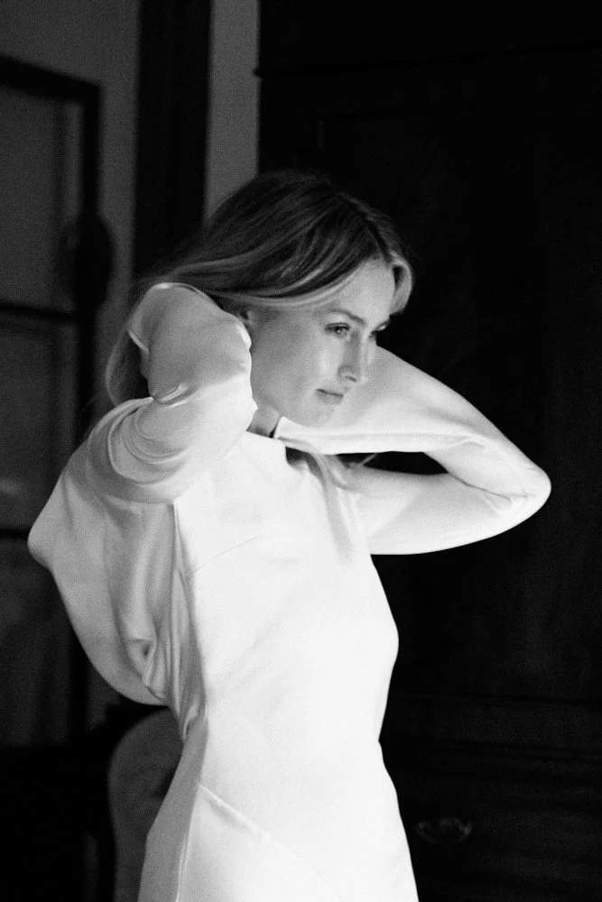 A woman in a long-sleeved white shirt is adjusting her hair, standing indoors near a dark wooden piece of furniture in a black-and-white photograph.
