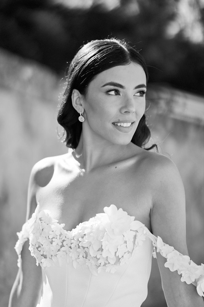 A woman in a wedding dress with floral embellishments, looking to the side outdoors.