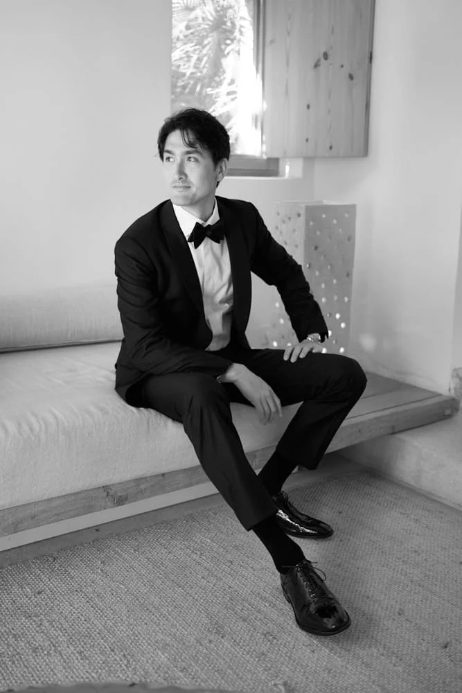 Man wearing a tuxedo sitting on a bench in a room with a window and wooden paneling, in black and white.