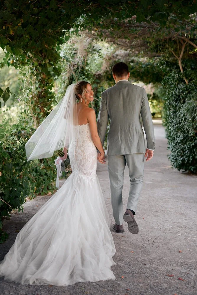Bride and groom walking hand in hand through a lush, green tunnel on their wedding day, with the bride wearing a white lace wedding gown and veil, and the groom in a light gray suit.