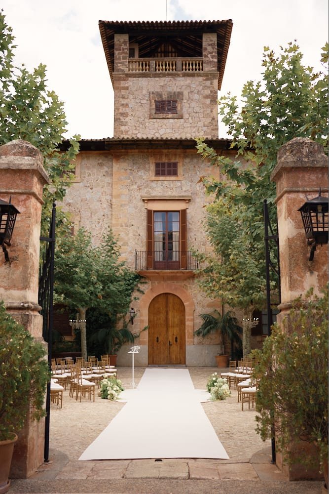 A rustic wedding venue featuring a stone building with a tower and wooden balcony, decorated with an aisle, chairs, and floral arrangements for a ceremony.