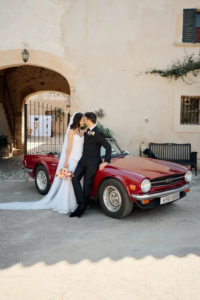 A bride and groom in wedding attire standing in front of a vintage red car, sharing a moment together outside a stone building with arched doorways and windows.