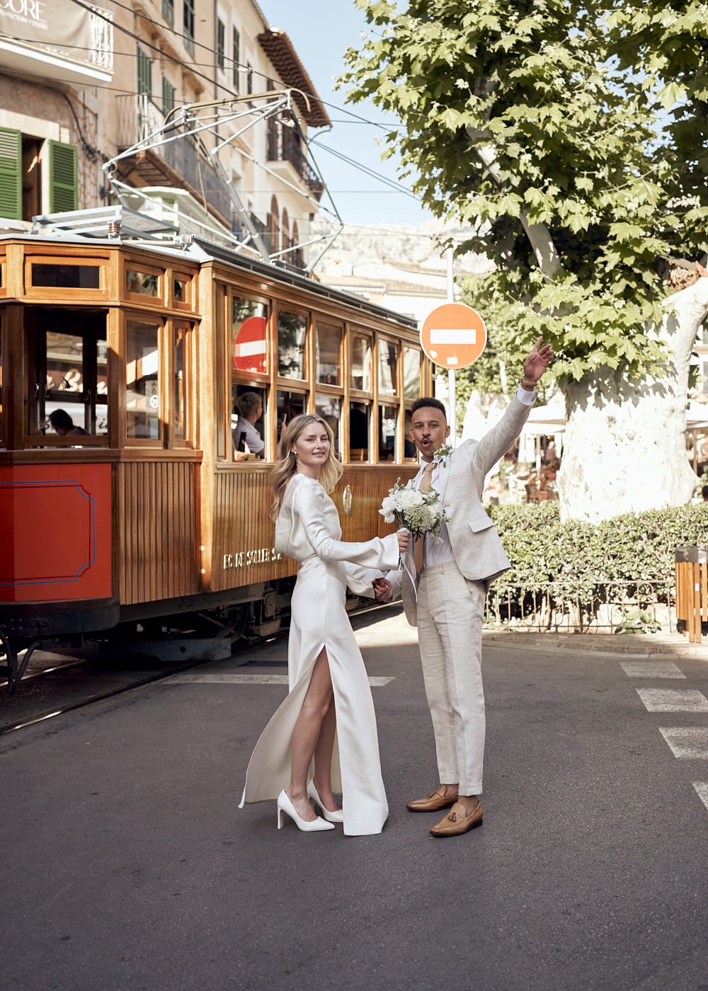 A bride and groom in formal attire pose happily on the street, with a vintage wooden tram and trees in the background. The groom raises his hand in celebration while the bride holds a bouquet of flowers.