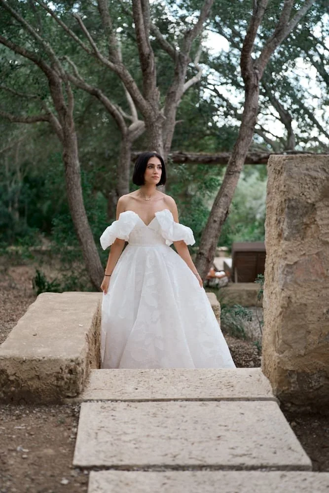 A woman in a white wedding dress with puffed sleeves standing outdoors between large stone blocks, with trees and greenery in the background.