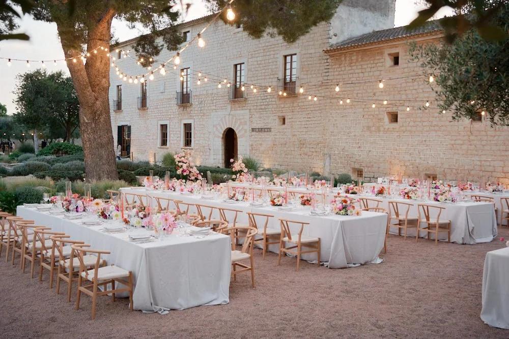 Outdoor wedding reception with long tables covered in white tablecloths, decorated with pink and white floral centerpieces, set up in front of a stone building with string lights hanging overhead and surrounding trees.