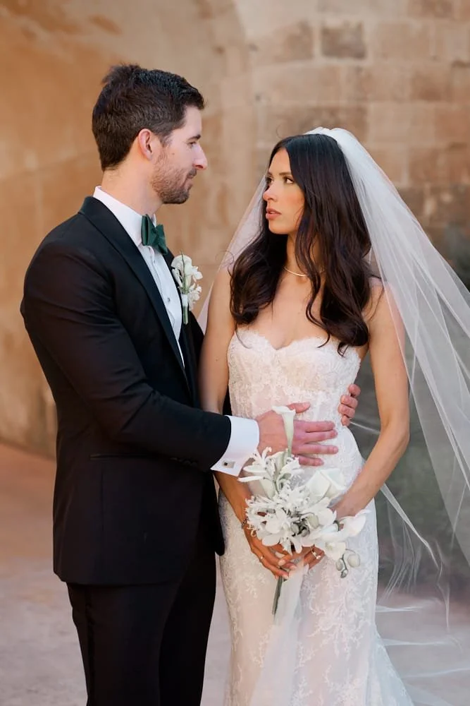 A bride and groom are standing close together during their wedding, with the groom in a black tuxedo and the bride in a white lace wedding dress and veil, holding a bouquet of white flowers.