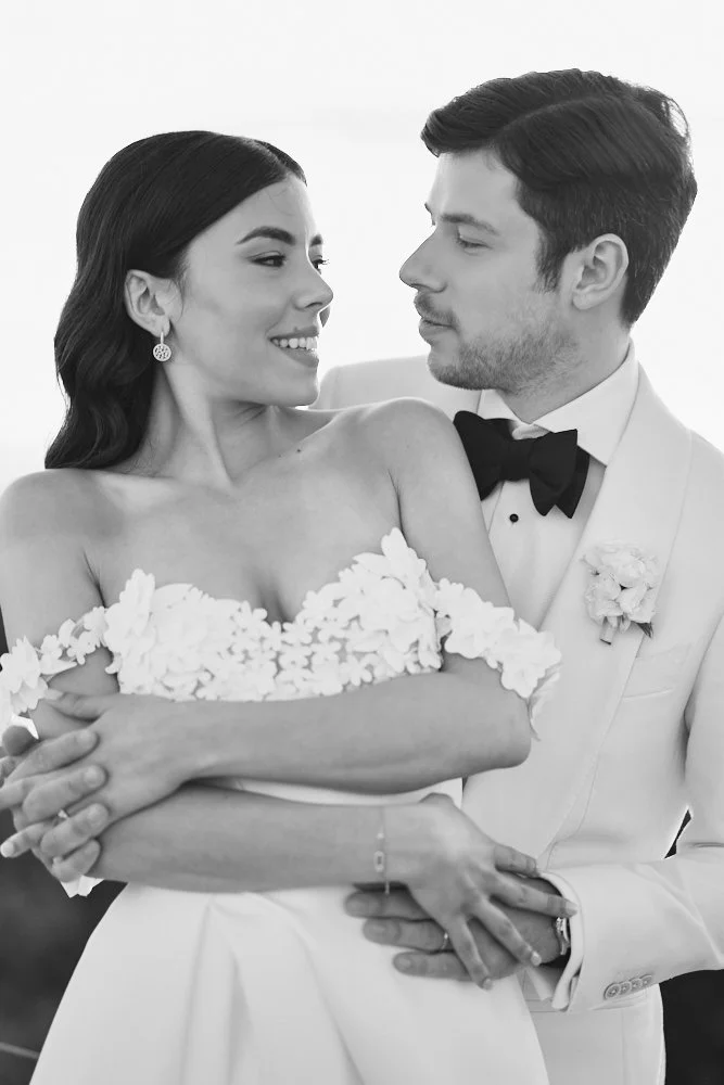Black and white photo of a bride and groom in wedding attire, sharing an intimate moment and smiling at each other.