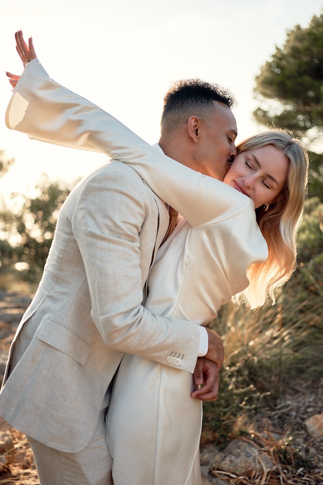 A couple in formal attire, a man and woman, sharing a kiss outdoors during sunset, with trees and natural landscape in the background.