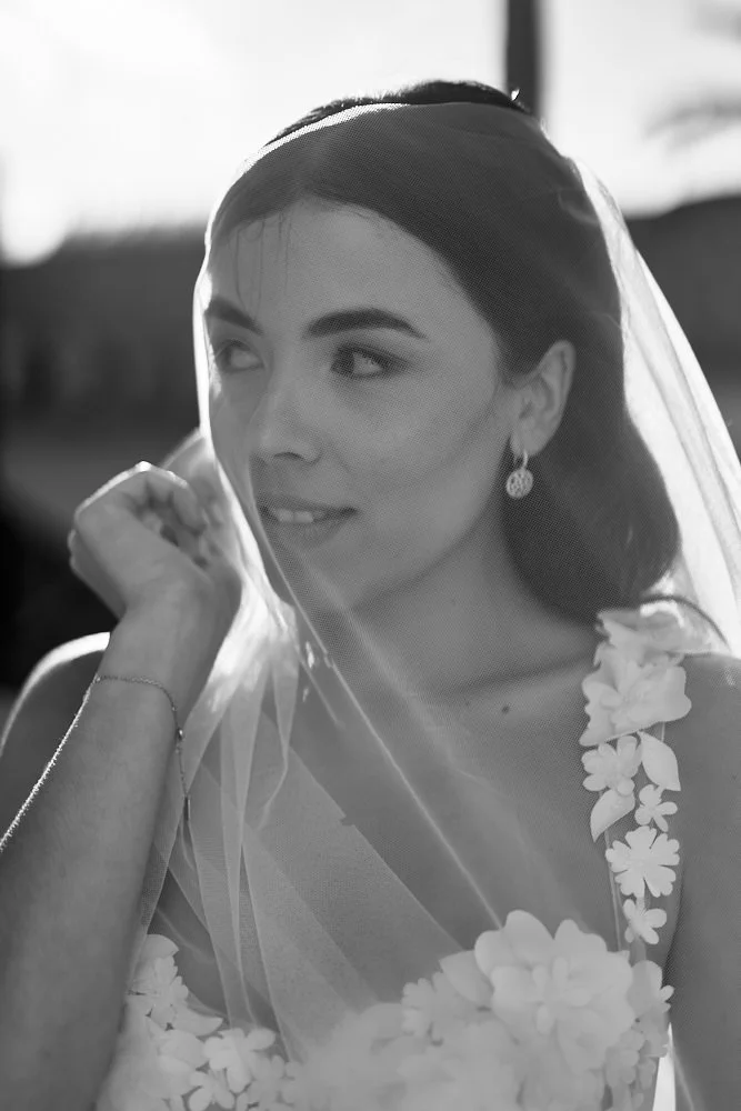 Black and white photo of a smiling bride holding her veil with her right hand, wearing floral dress and earrings, outdoors with sunlight.