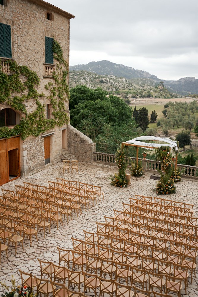 Outdoor wedding ceremony setup on a stone-paved courtyard with rows of wooden chairs, an arch decorated with flowers, and a scenic mountain view in the background.