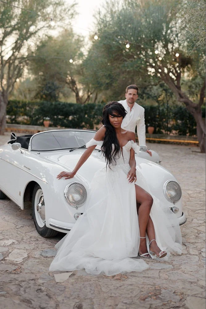 A woman in a white wedding dress with a thigh-high slit and heels, sitting on the front of a white vintage convertible car with a man standing behind her, outdoors in a park-like setting with trees and string lights.